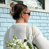 Image of girl wearing our blue and white check daydream classic claw in her hair. She is wearing a white sweater and tortoiseshell sunglasses. She is carrying a white cane bag with white flowers in the bag.