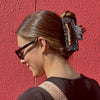 Image of girl wearing our classic extra large claw in domino. She is against a red wall and is wearing a black top with black sunglasses. 