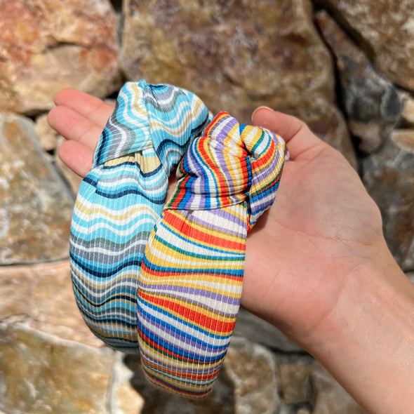 Picture of ladies hand holding two headbands.  One is in horizontal colours of blues, white, greys and mustard.  The other is multicoloured in horizontal colours of red, blue, green, yellows and greys.  Both have knotted tops.